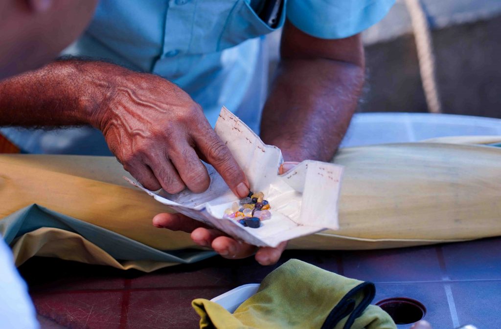 A Sri Lankan gem trader carefully inspects a collection of raw, uncut gemstones wrapped in traditional paper. 